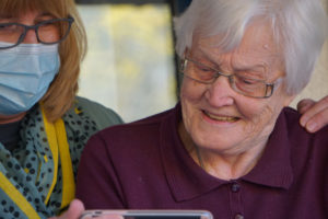 A carer in an aged care environment helps an elderly lady with technology. Both are wearing glasses and are working together. This is to demonstrate carer self-care tips and how helping you is essential to helping others.