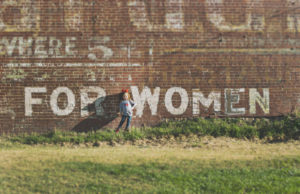 a young girl stands against a brick wall that reads "for women"