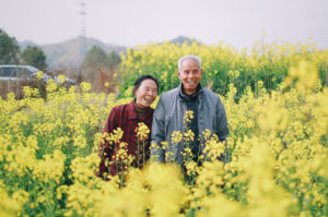 an elderly asian couple laugh in a crop of golden flowers