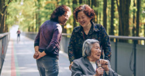 An elderly Asian lady is pushed in a park in a wheelchair while two other people talk happily together