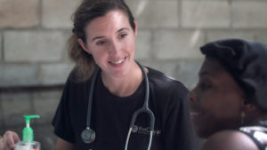 a woman in scrubs with a stethoscope talks to an elderly person of colour